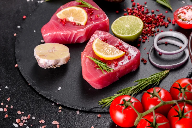 Raw tuna fillets with lemon slices, herbs, garlic, and tomatoes on a black slate