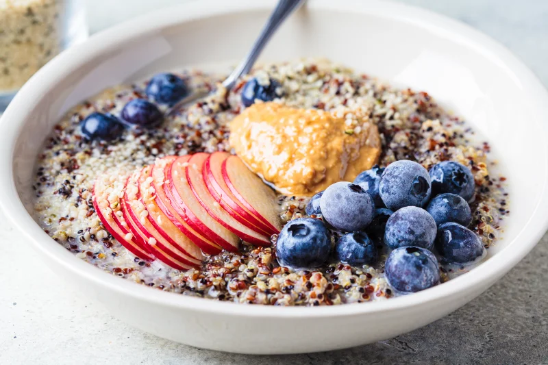 Quinoa Porridge with Fruits and Peanut Butter Bowl of quinoa porridge topped with blueberries, apple slices, and peanut butter for plant protein and energy.