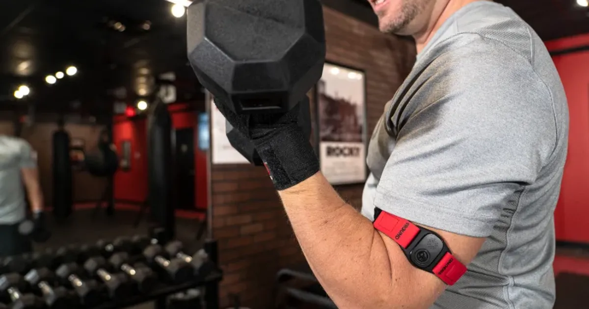 Man lifting dumbbell while wearing 9Round PULSE heart rate armband in gym.