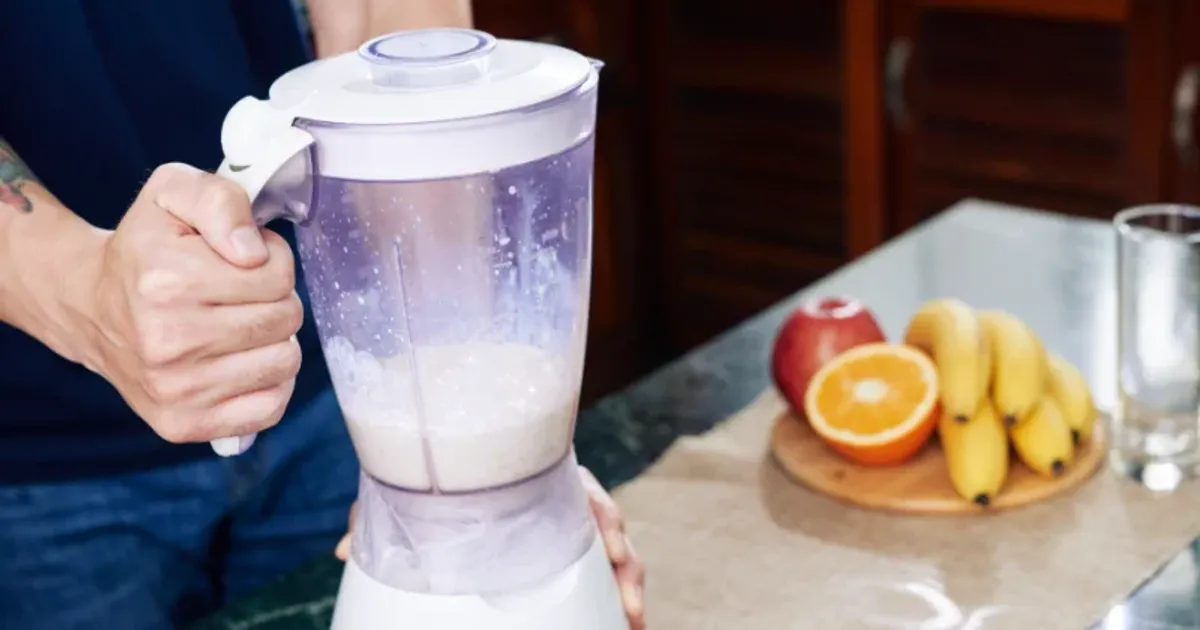 Man preparing a protein smoothie in a blender with bananas and orange on the counter for post-workout recovery.
