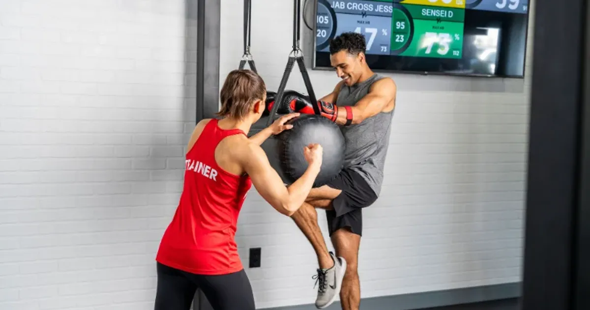 Trainer coaching man performing knee strikes on heavy bag at 9Round with heart rate feedback screen