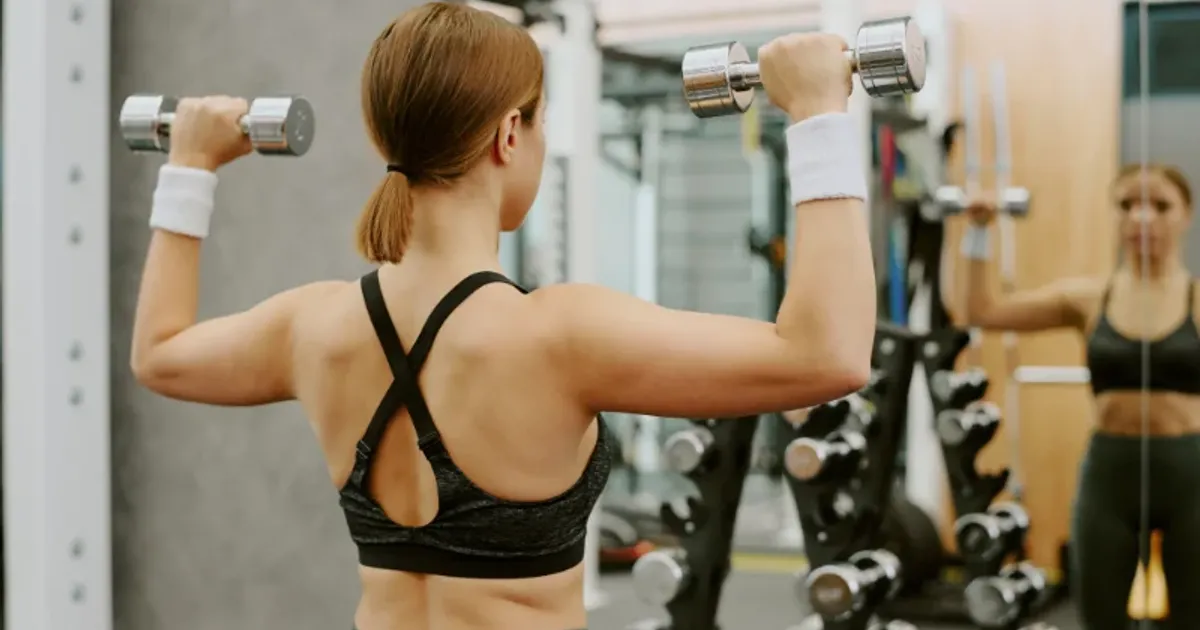 Young woman lifting dumbbells for shoulder workout while looking in gym mirror.