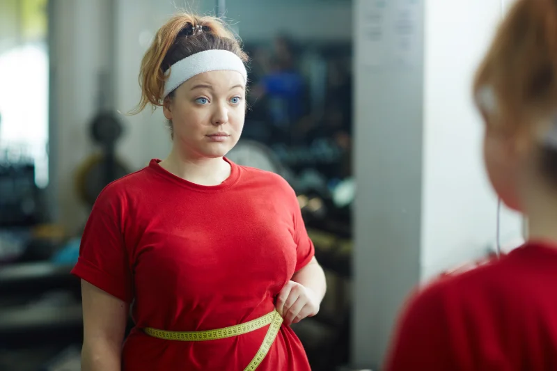 Tracking Fitness Progress During 30-Minute Daily Workouts Young woman in red workout shirt measuring waistline with tape in gym mirror.