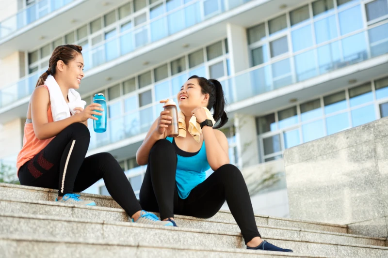Two women smiling and hydrating after an outdoor workout on building steps