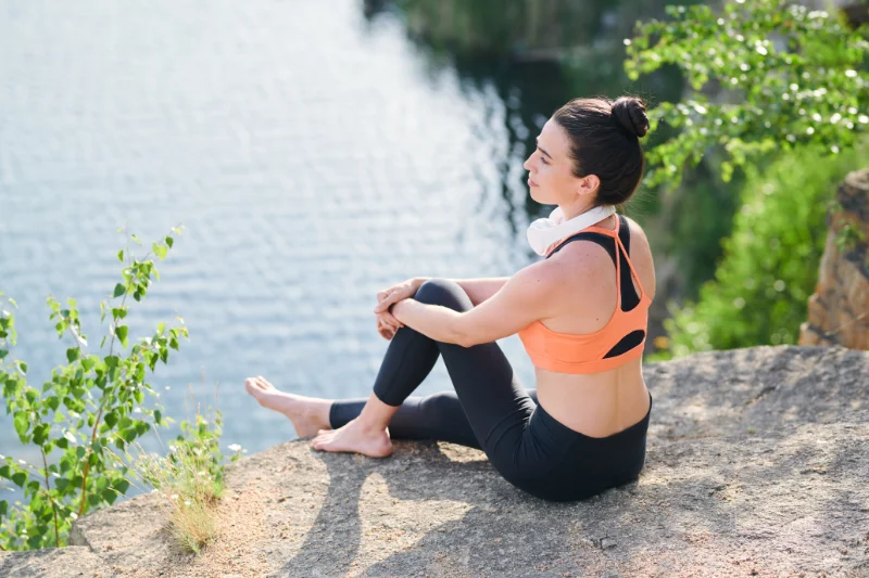 Post-Workout Recovery by the Lake Woman in activewear resting by a lakeside with headphones around her neck.