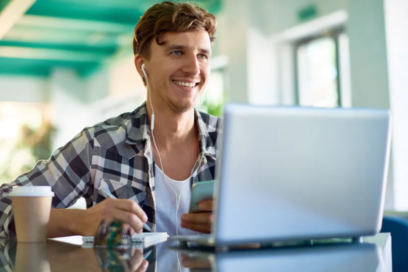 Smiling young man studying at laptop with headphones and coffee cup.
