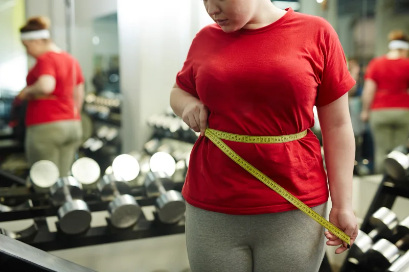 Woman in a red shirt measuring her waistline with a tape at the gym after HIIT training